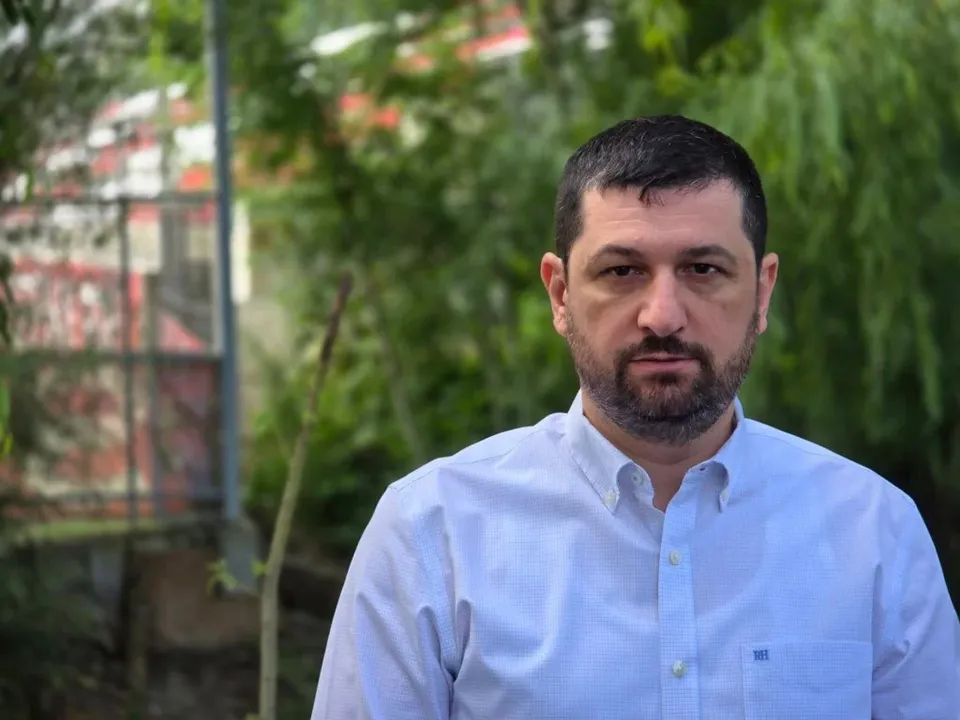 Un bărbat cu barbă și păr scurt, purtând o cămașă albă, stă în fața unui fundal cu vegetație verde și o clădire difuză.