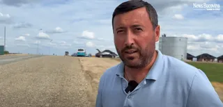 Un bărbat cu barbă, îmbrăcat într-un tricou polo albastru deschis, stă în prim-plan, cu un drum de pământ și vehicule în fundal, sub un cer noros.