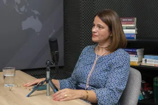 O femeie cu părul scurt și brunet poartă o bluză albastră cu modele geometrice și un colier de perle, stând la o masă de lemn.