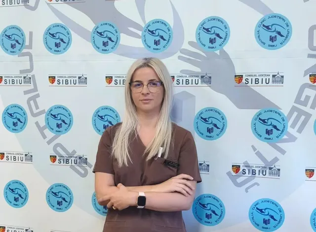 O femeie cu părul blond și ochelari stă cu brațele încrucișate în fața unui fundal alb cu diverse sigilii și logouri circulare albastre.