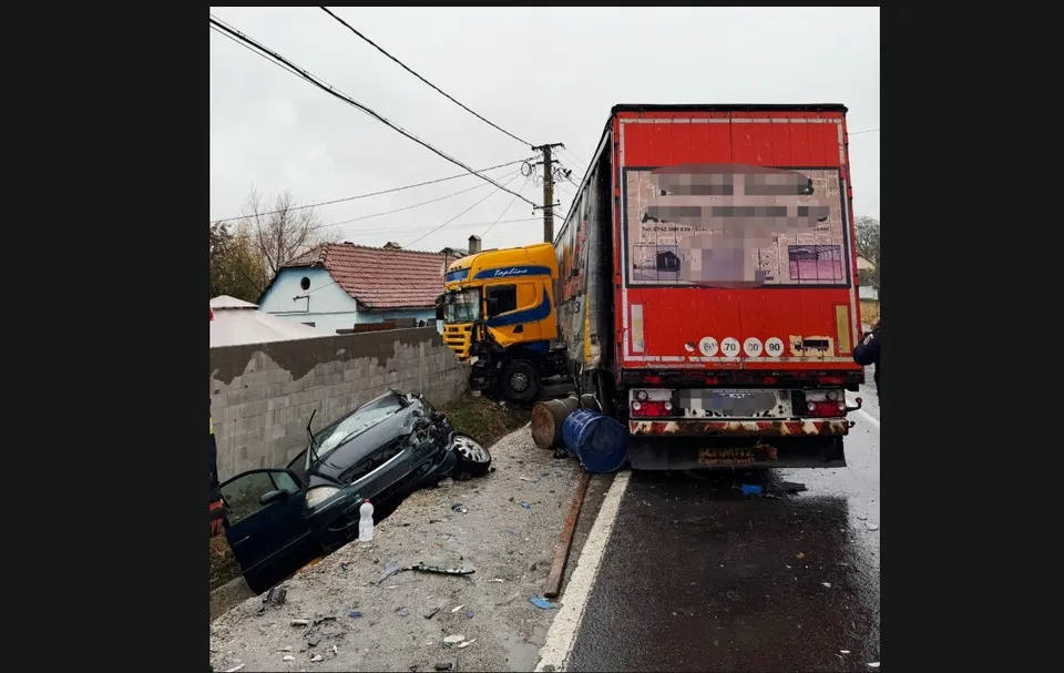 Un autocamion galben și roșu este oprit pe marginea unui drum lângă un autoturism negru avariat și resturi împrăștiate pe sol.