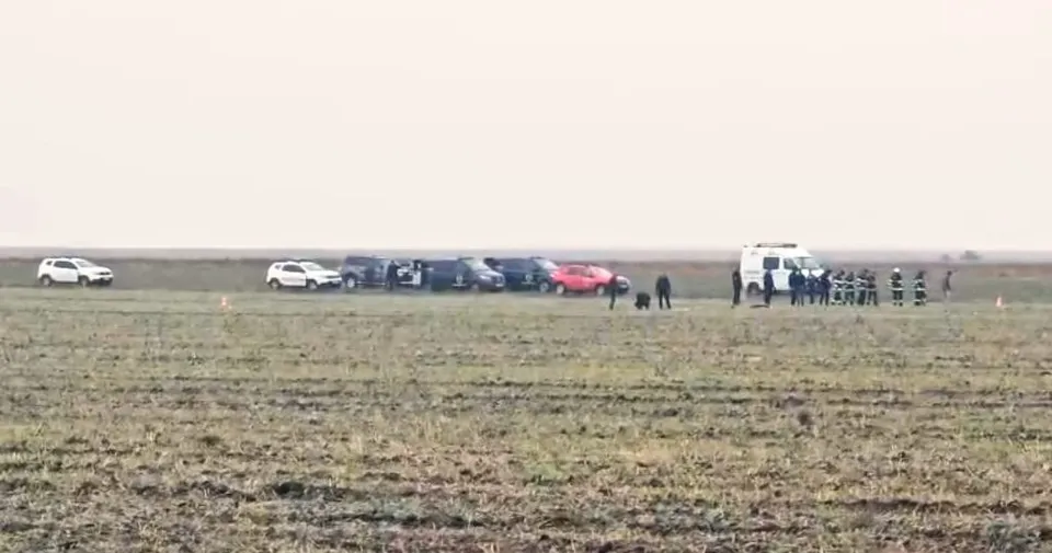 Mai multe mașini și un van alb sunt parcate într-un câmp deschis lângă un grup de persoane îmbrăcate în uniforme în depărtare.