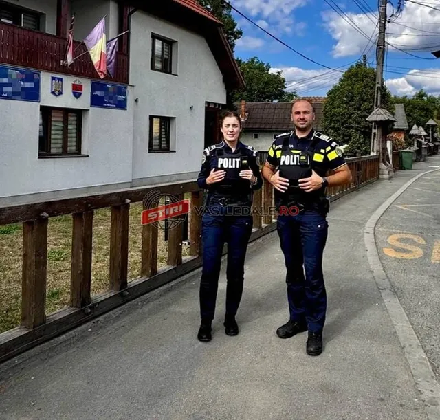 Doi ofițeri de poliție, un bărbat și o femeie, stau în fața unei clădiri, pe un trotuar, cu un gard de lemn în prim-plan.