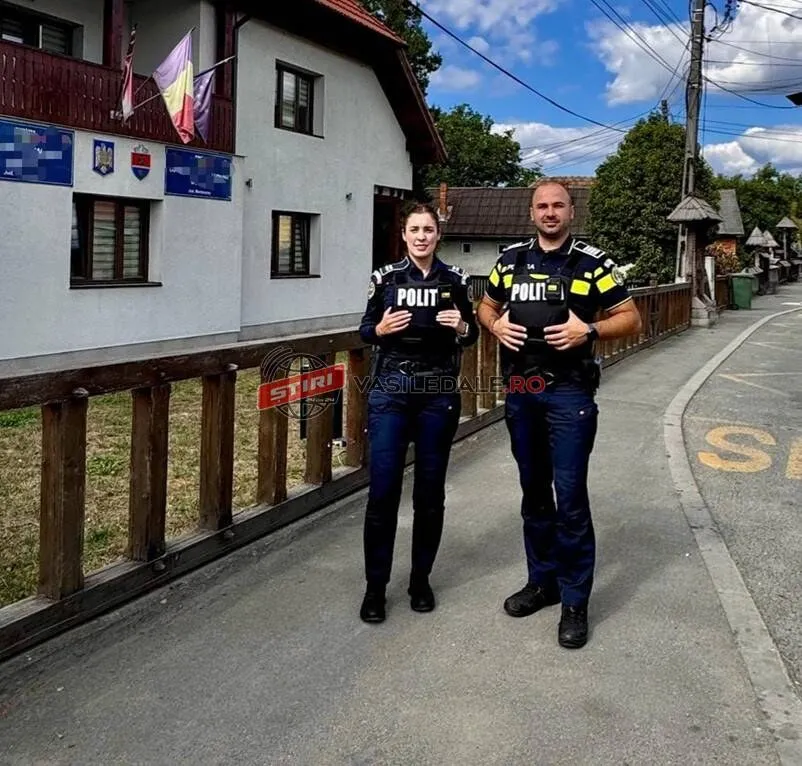 Doi ofițeri de poliție, un bărbat și o femeie, stau în fața unei clădiri, pe un trotuar, cu un gard de lemn în prim-plan.