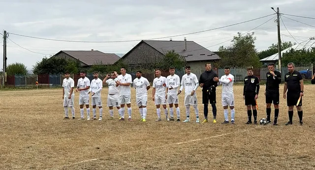 O echipă de fotbaliști în echipament alb stă în linie pe un teren de fotbal, alături de arbitri și adversari, sub un cer înnorat.