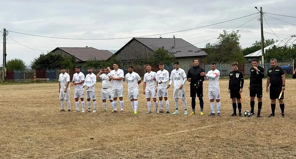 O echipă de fotbaliști în echipament alb stă în linie pe un teren de fotbal, alături de arbitri și adversari, sub un cer înnorat.