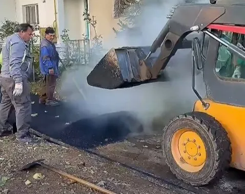 Un utilaj galben descarcă material negru pe o suprafață asfaltată în prezența a doi bărbați îmbrăcați în haine de lucru.
