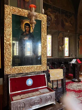 O icoană înrămată aurit este suspendată deasupra unui relicviar metalic cu interiorul căptușit în catifea roșie, într-un interior de biserică decorat.