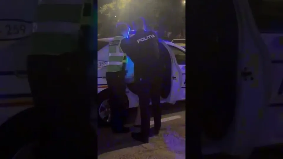 Un agent de poliție cu inscripția POLITIA pe uniformă stă lângă o mașină albă în timpul nopții.