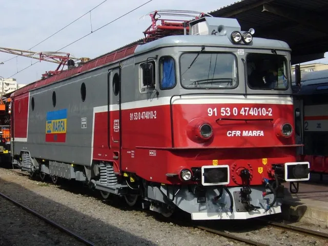 O locomotivă gri și roșie cu inscripția CFR MARFA stă pe o cale ferată lângă un acoperiș de stație.