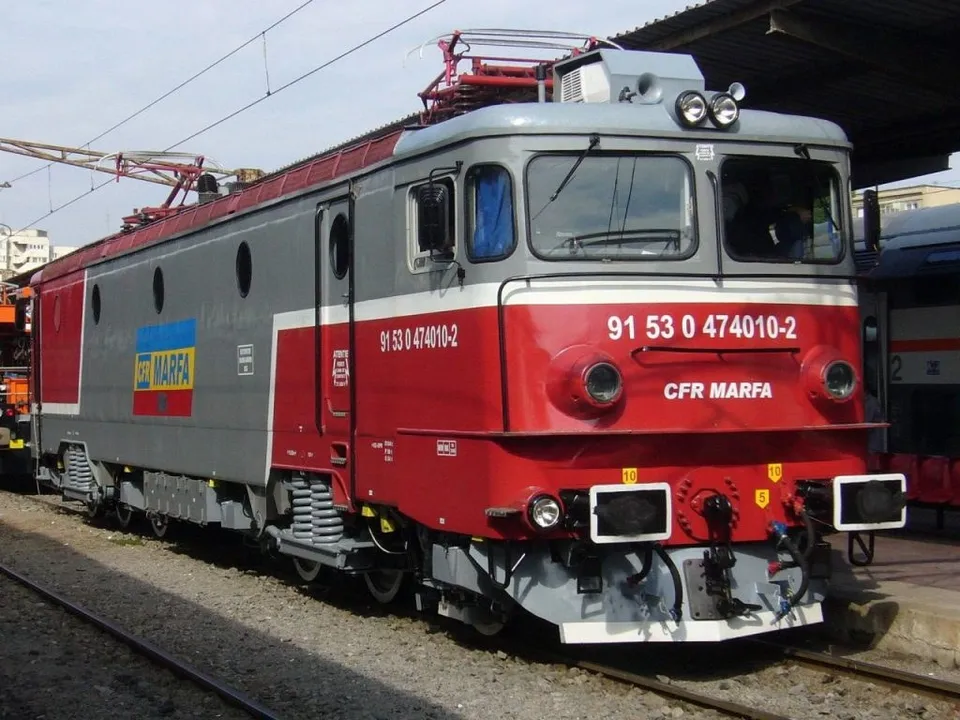 O locomotivă gri și roșie cu inscripția CFR MARFA stă pe o cale ferată lângă un acoperiș de stație.
