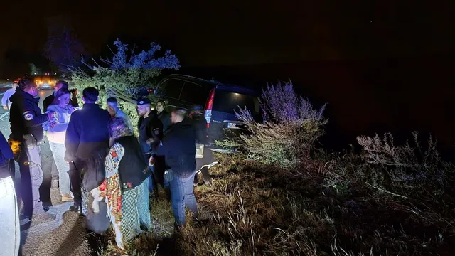 Mai mulți oameni stau pe marginea unui drum lângă un vehicul înfundat într-o vegetație densă, sub lumina reflectoarelor, noaptea.