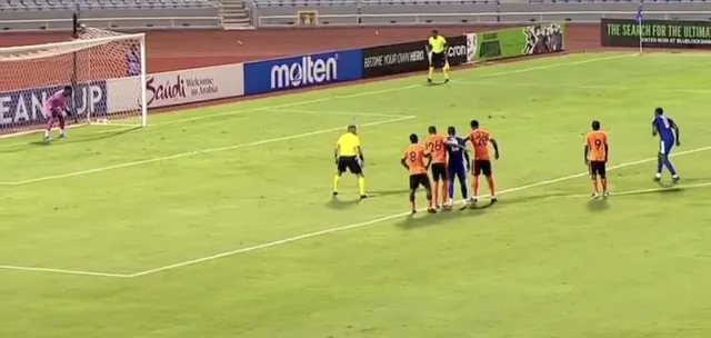 Câțiva jucători de fotbal stau pe terenul de joc lângă o linie albă, în timp ce un portar se află în fața porții.
