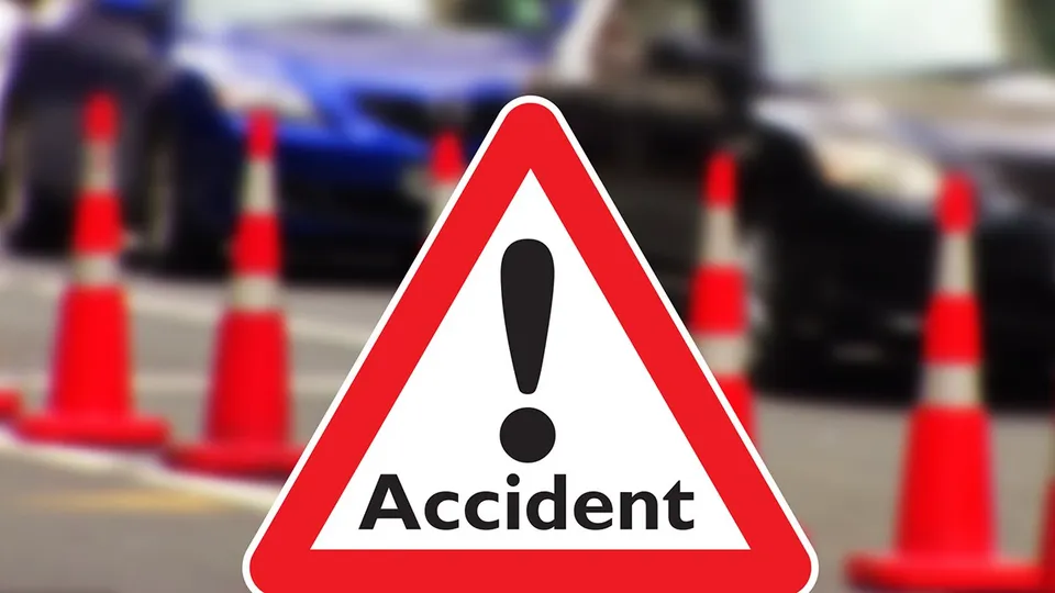 Un semn de circulație triunghiular cu margini roșii și un semn exclamării negru este plasat în prim-plan, în fața unor conuri de trafic și mașini.