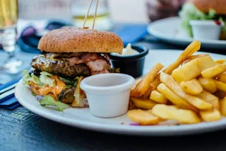 Un burger cu bacon și salată este servit pe o farfurie albă alături de cartofi prăjiți și un bol mic cu sos.