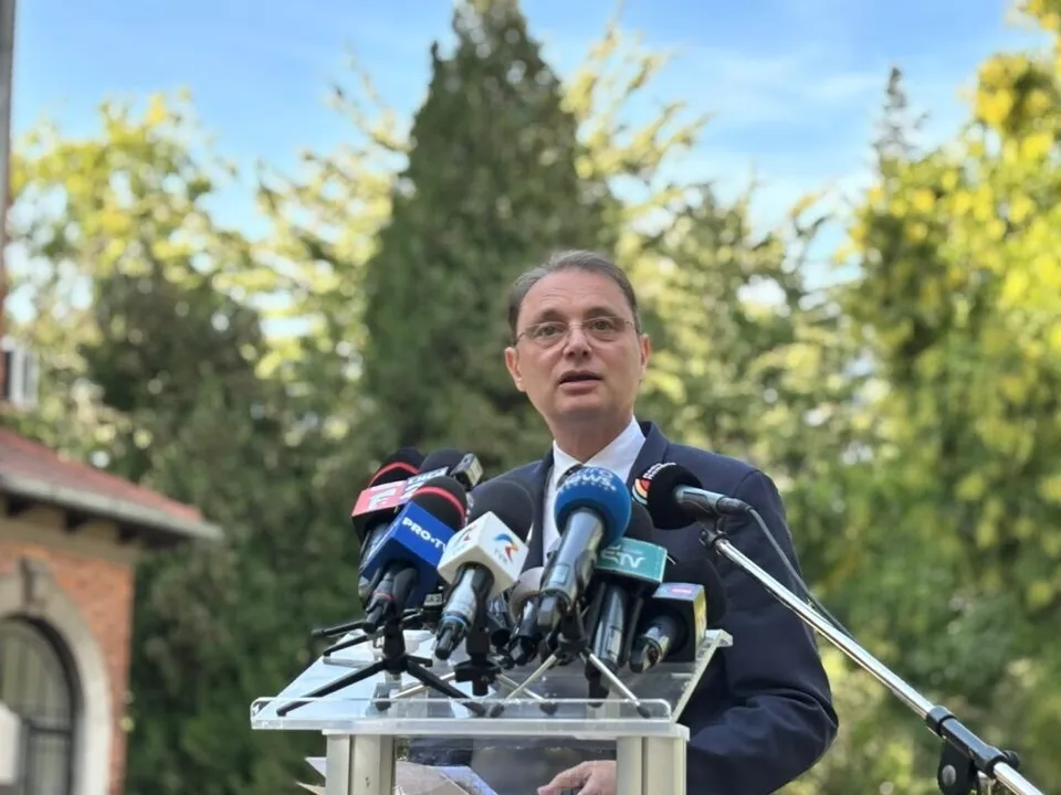 Un bărbat cu ochelari și costum închis la culoare vorbește în fața mai multor microfoane dispuse pe un suport metalic.
