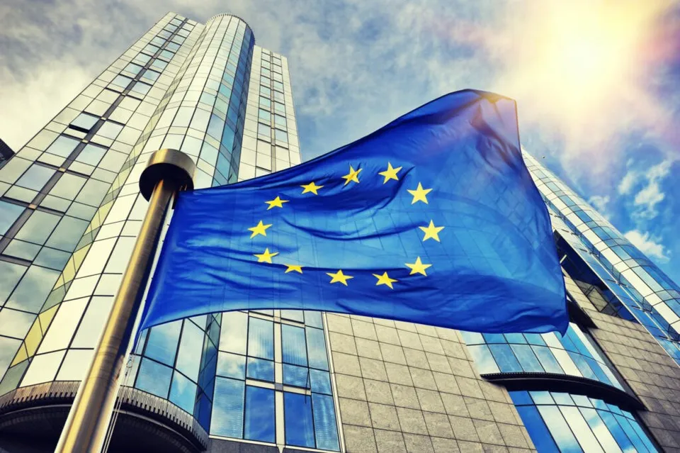 O steag al Uniunii Europene flutură pe un stâlp în fața unei clădiri moderne cu fațadă de sticlă sub un cer însorit.