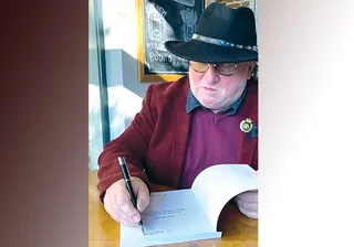 Un bărbat cu ochelari și pălărie neagră scrie cu un pix într-o carte deschisă aflată pe o masă de lemn.