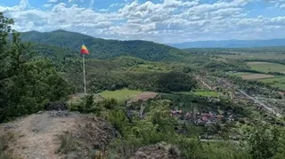 O vedere panoramică de la înălțime asupra unui peisaj montan cu dealuri verzi, păduri și un sat cu case mici în vale sub un cer noros.