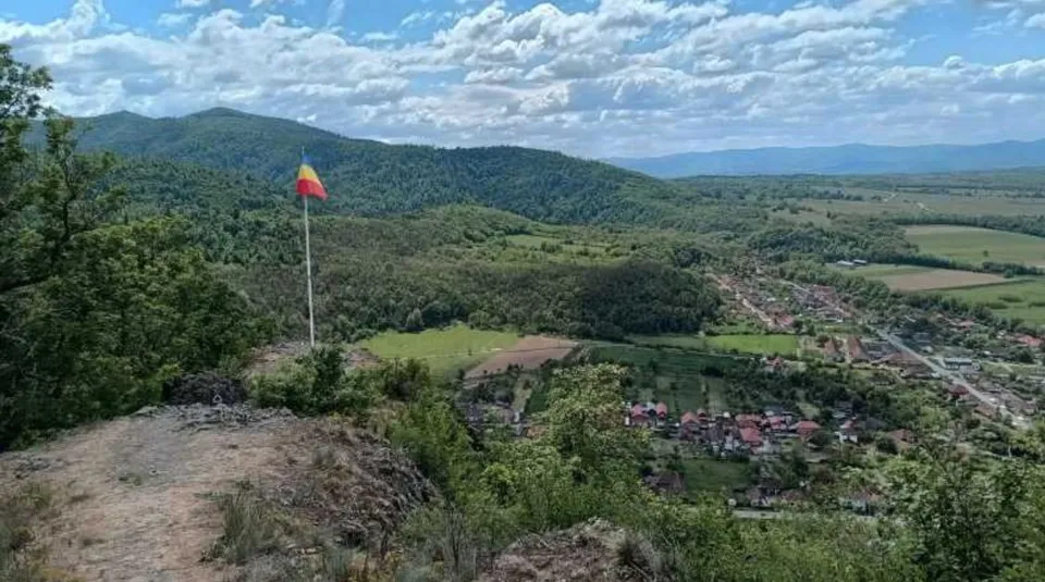 O vedere panoramică de la înălțime asupra unui peisaj montan cu dealuri verzi, păduri și un sat cu case mici în vale sub un cer noros.
