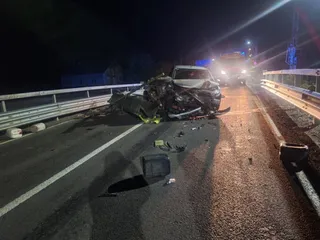Un autoturism distrus stă pe marginea unei drumuri asfaltate lângă o barieră metalică, în timp ce un vehicul de servicii cu lumini aprinse se apropie.