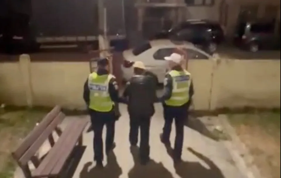 Trei persoane, dintre care două poartă veste reflectorizante, merg pe un trotuar lângă o bancă de lemn și mașini parcate.