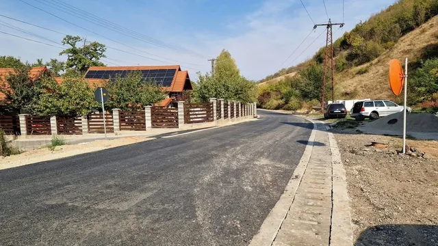 O porțiune de drum asfaltat cu borduri de beton este încadrată de un gard de lemn, vegetație și un stâlp de înaltă tensiune.