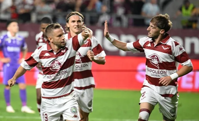 Câțiva jucători de fotbal în echipamente de culoare albă și roșie sărbătoresc pe un teren de fotbal sub luminile stadionului.
