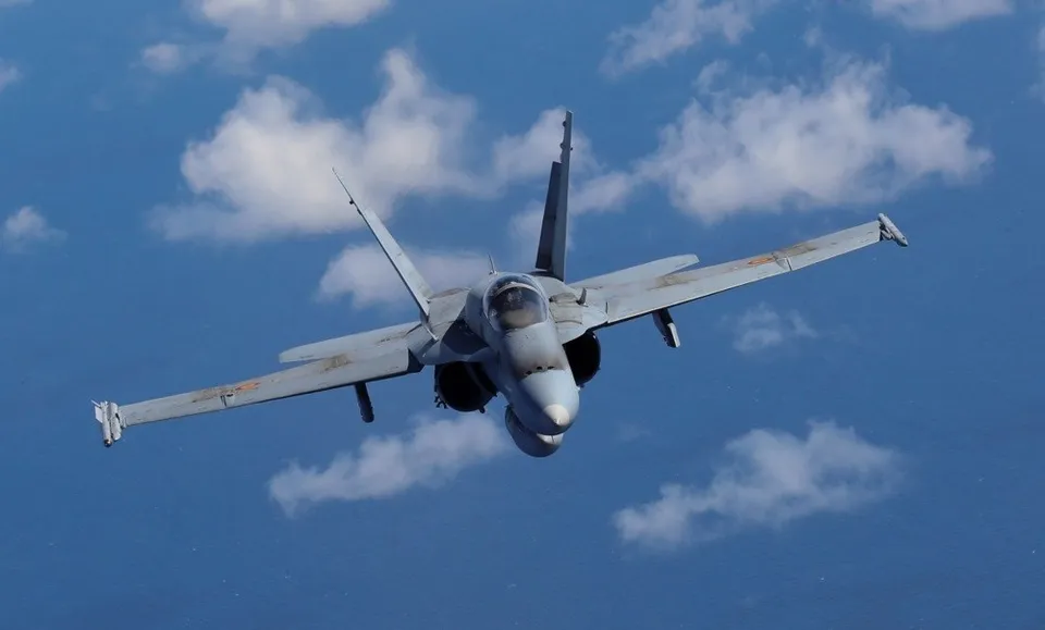 Un avion de luptă gri zboară pe un cer albastru cu nori albi dispersați.