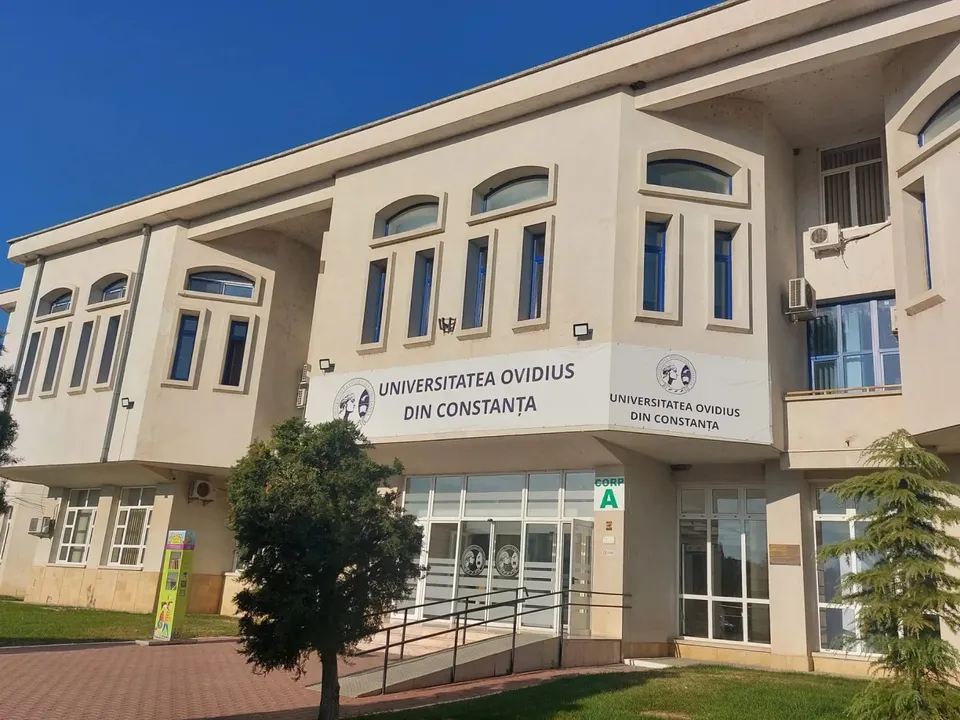 O clădire cu fațadă albă prezintă ferestre înalte, un panou cu textul „UNIVERSITATEA OVIDIUS DIN CONSTANȚA” și o intrare cu uși de sticlă.