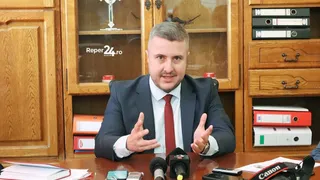 Un bărbat cu cămașă albă, cravată roșie și sacou închis la culoare stă la o masă de lemn în fața unor dulapuri cu obiecte și dosare.
