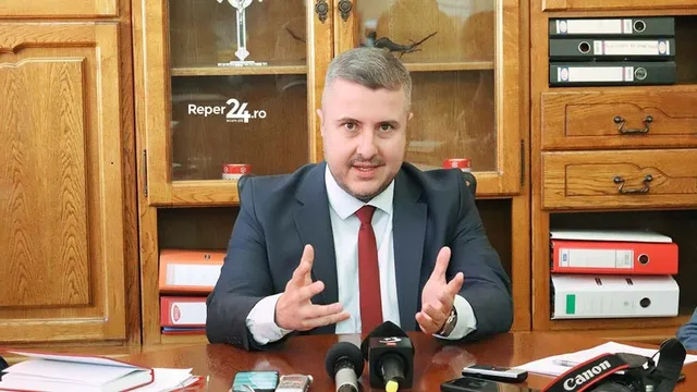 Un bărbat cu cămașă albă, cravată roșie și sacou închis la culoare stă la o masă de lemn în fața unor dulapuri cu obiecte și dosare.