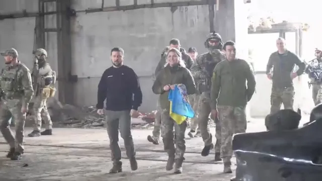 Un grup de persoane în haine militare și civile merge printr-un spațiu deschis și luminos cu pereți de beton.