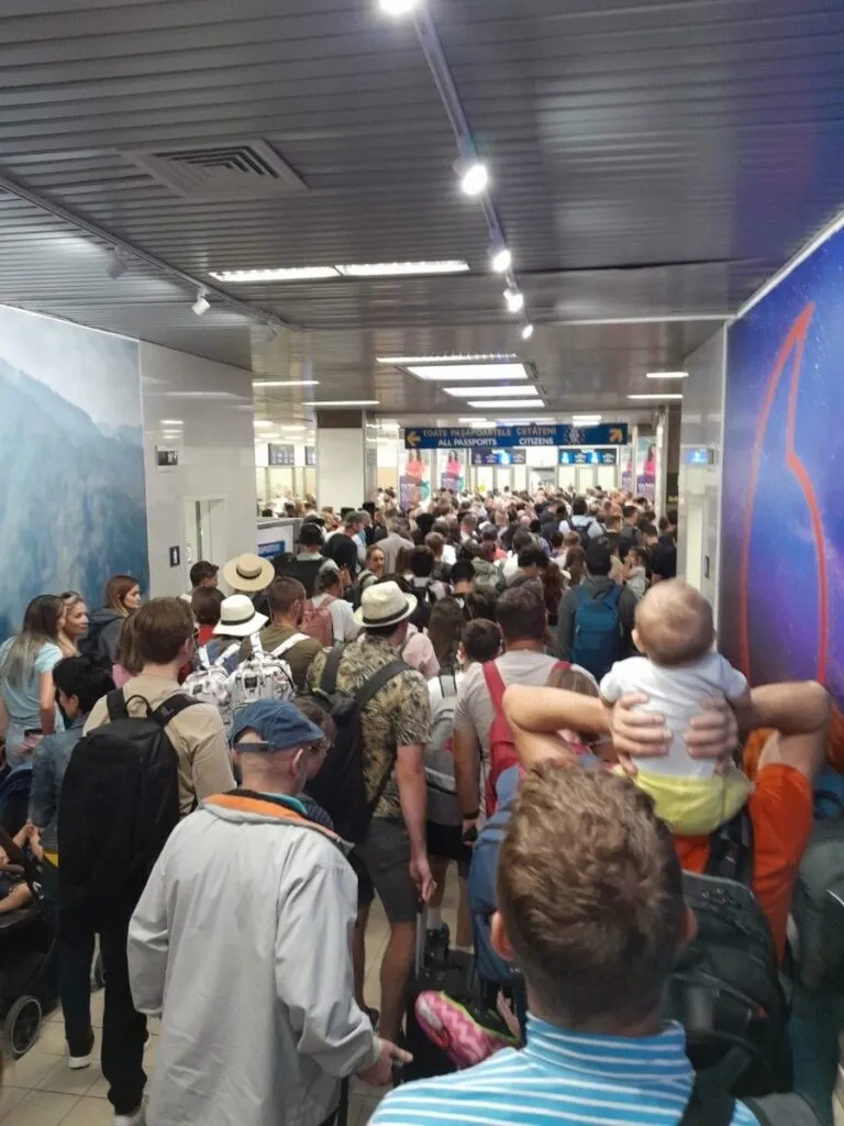 O mulțime de oameni stă în picioare într-un coridor cu tavan înălțat și lumini alungite, purtând haine casual și rucsacuri.