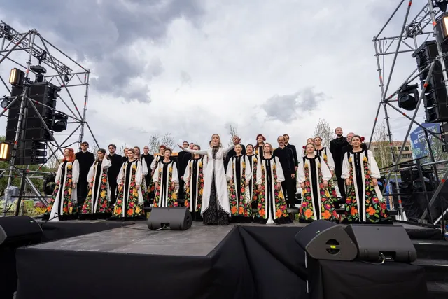 Un grup mare de coriști în haine tradiționale albe cu motive florale stă pe o scenă sub un cer înnorat.