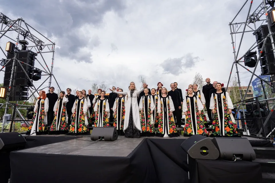 Un grup mare de coriști în haine tradiționale albe cu motive florale stă pe o scenă sub un cer înnorat.
