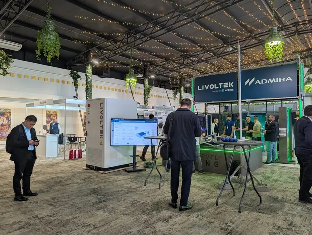 Oamenii vizitează un stand de expoziție cu panouri informative și echipamente tehnice sub un plafon cu structură metalică și lumini decorative.