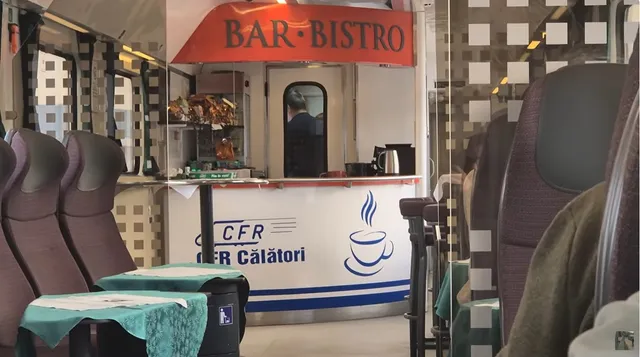 Un bar-bistro situat într-un compartiment de tren, cu scaune cu spătar înalt și mese mici acoperite cu fețe de masă verzi.