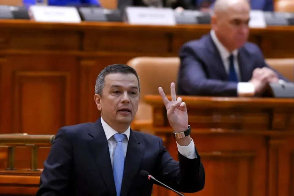 Un bărbat într-un costum închis și cravată albastră stă în fața unui microfon, ridicând mâna stângă cu degetul arătător și mijlociu întindeți.
