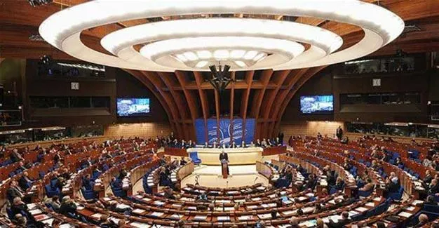 O sală de conferințe mare cu scaune dispuse în semicerc sub un plafon circular iluminat, având un podium central unde stă o persoană.