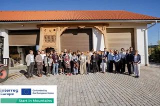 Un grup de persoane stă în rând în fața unei clădiri albe cu uși de garaj maronii și un acoperiș de țiglă portocalie.