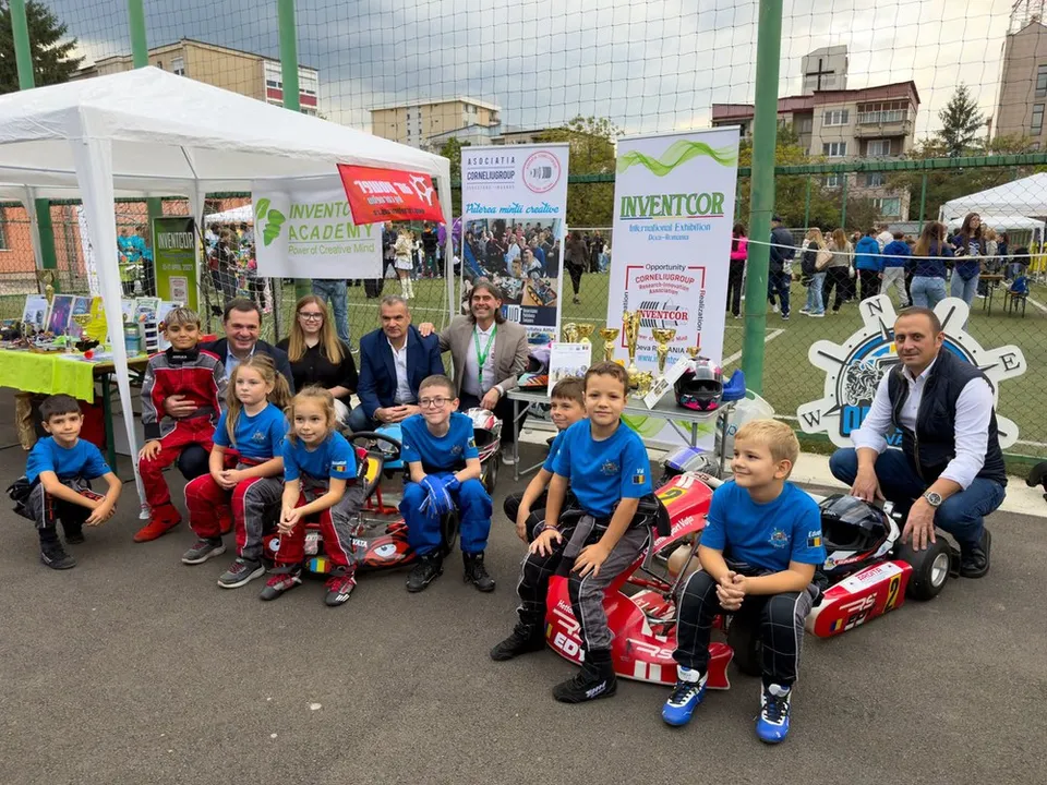 Un grup de copii în tricouri albastre și adulți stau poziționați în fața unor corturi și panouri publicitare pe un teren de sport.