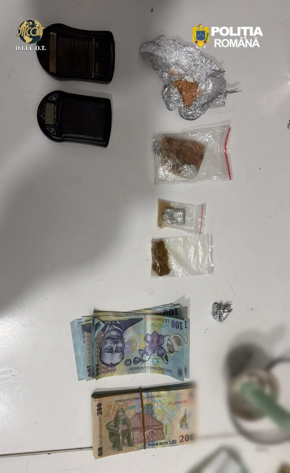 Mai multe plicuri mici transparente conțin substanțe maronii, alături de câteva bancnote și două dispozitive electronice negre pe o suprafață albă.