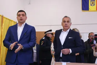 Un bărbat într-un costum albastru și unul într-un costum închis stau în picioare într-o încăpere cu un oficial uniformizat în fundal.