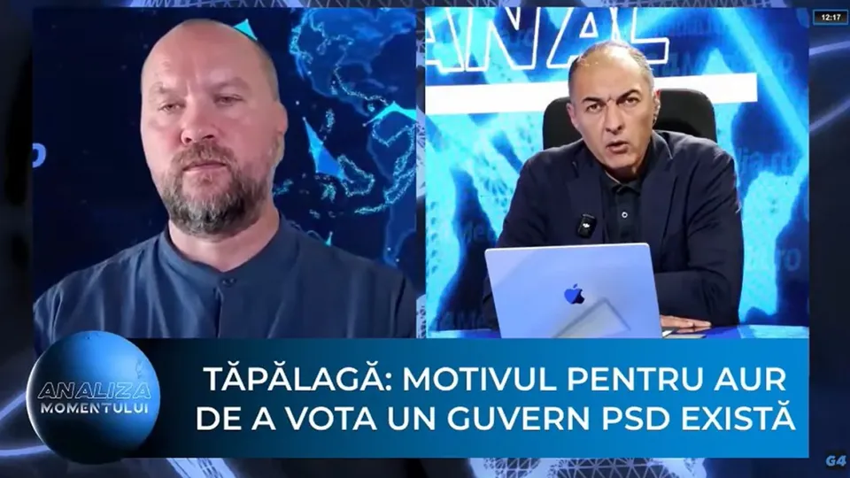 Un bărbat cu barbă poartă o cămașă închisă la culoare, în timp ce un alt bărbat stă la un birou lângă un laptop deschis.