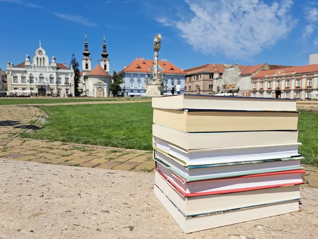 O stivă de cărți colorate este așezată pe o suprafață plană, având în fundal o piață cu clădiri istorice și un cer senin.