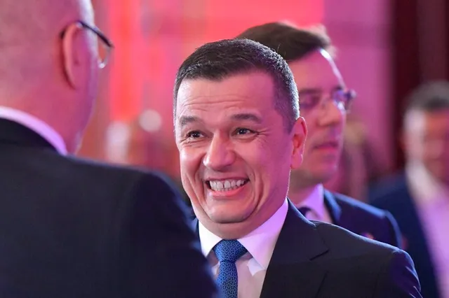 Un bărbat zâmbind într-un costum închis cu cămașă albă și cravată albastră este înconjurat de alți bărbați îmbrăcați în costume.