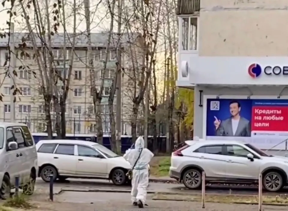 O persoană purtând un costum de protecție alb merge pe un trotuar lângă mașini parcate și clădiri în fața unor copaci.