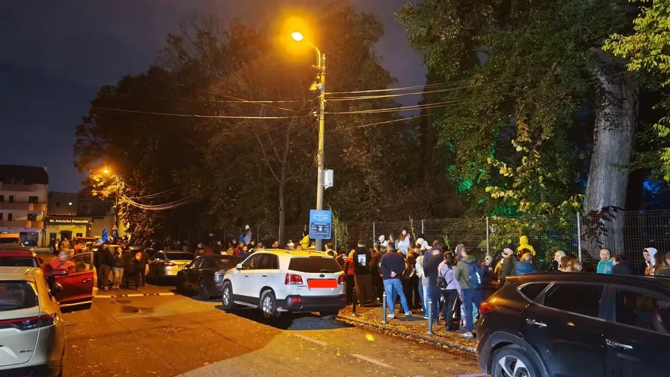 O mulțime de oameni stă pe un trotuar lângă mașini parcate sub un stâlp de iluminat public într-o zonă cu copaci noaptea.