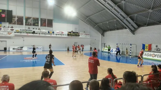 Câțiva jucători de handbal în echipamente sportive participă la un meci pe un teren de culoare albastră și maro într-o sală de sport închisă.